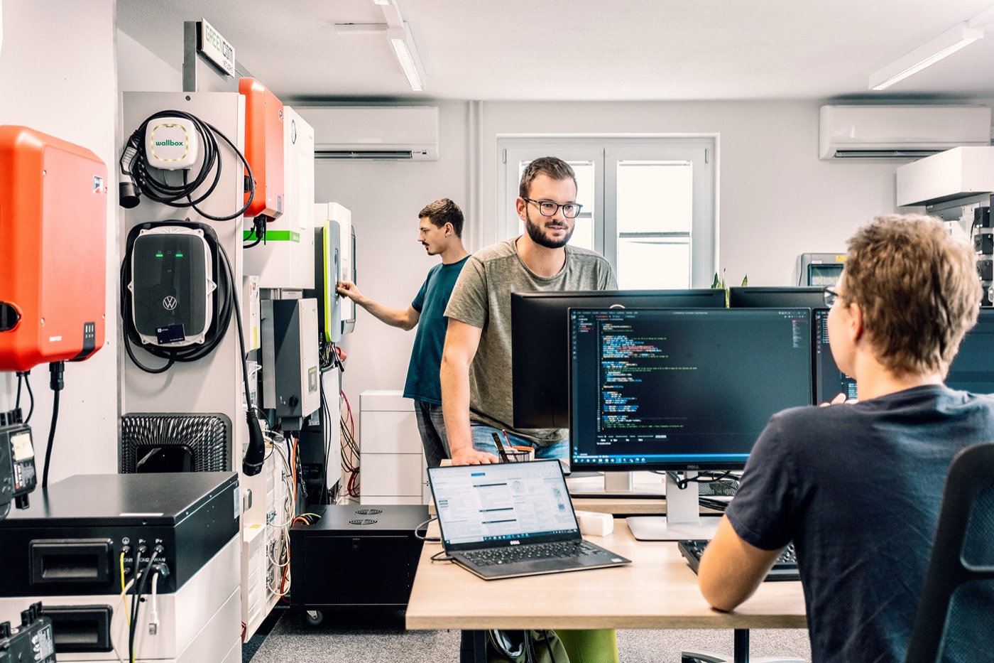 An engineer reading protocol traces on screen while a colleague operates a wallbox and inverters on the lab wall